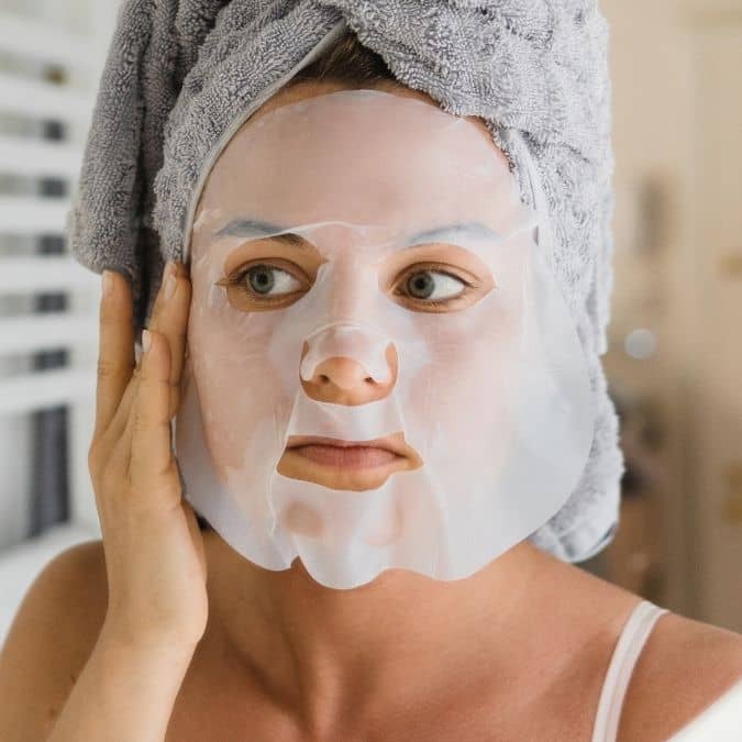 Woman with a gray towel wrapped around her head wearing a white sheet face mask while gently touching her cheek, promoting skincare and wellness with Moodmask - just let it glow | Patchology.