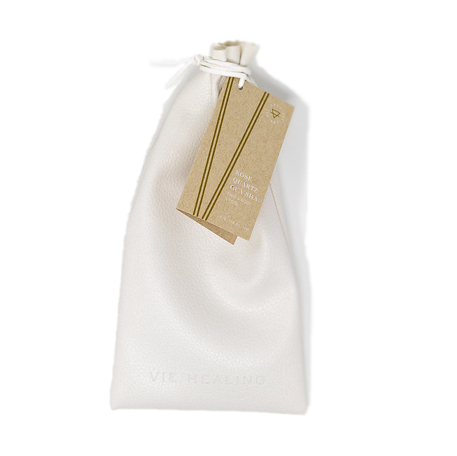 White textured drawstring pouch with a brown tag attached, containing the Rose Quartz Gua Sha | Vie Healing skincare tool inside for wellness and facial massage.