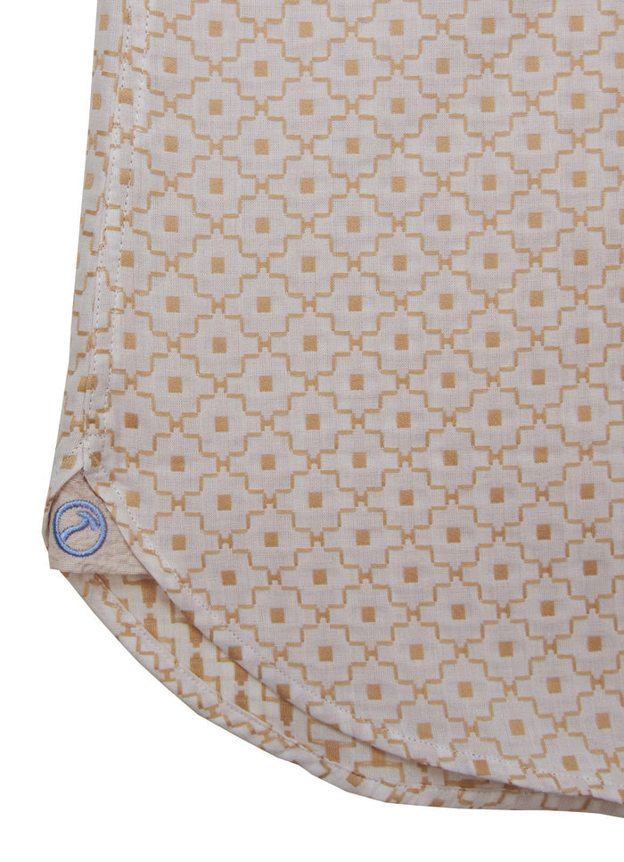 Close-up of the bottom corner of an Alimagnet beige cotton shirt featuring a geometric square neutral pattern, rounded hem, visible stitching, and a small blue logo tag.