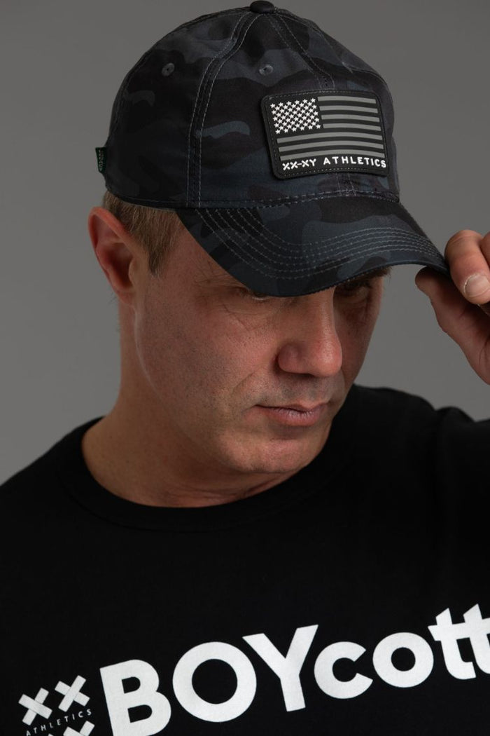 Man wearing a black cap with an American flag design and 'XX Athletics' text, against a gray background. || Black Camo