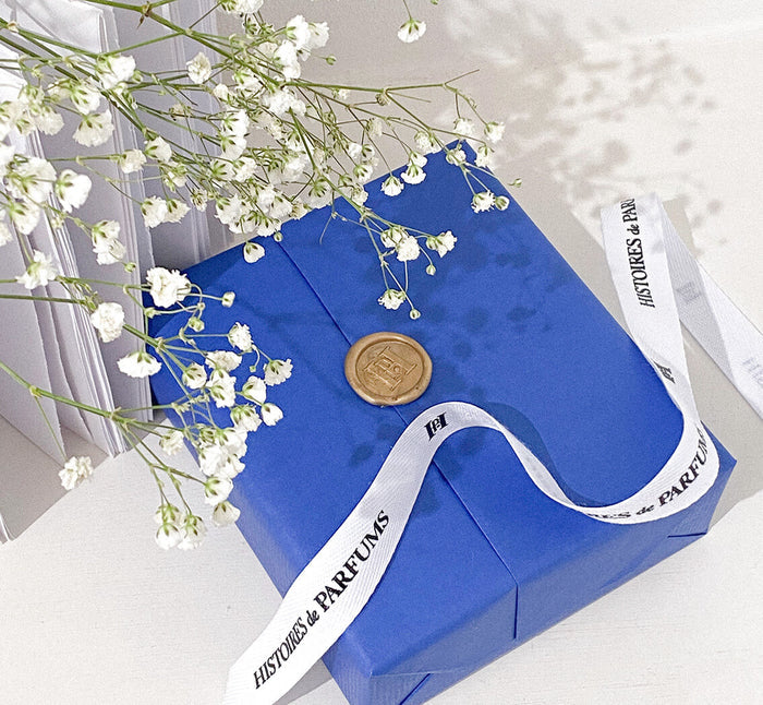A blue gift with a white ribbon and a gold-colored seal, resting on a surface with a white flower in the background.