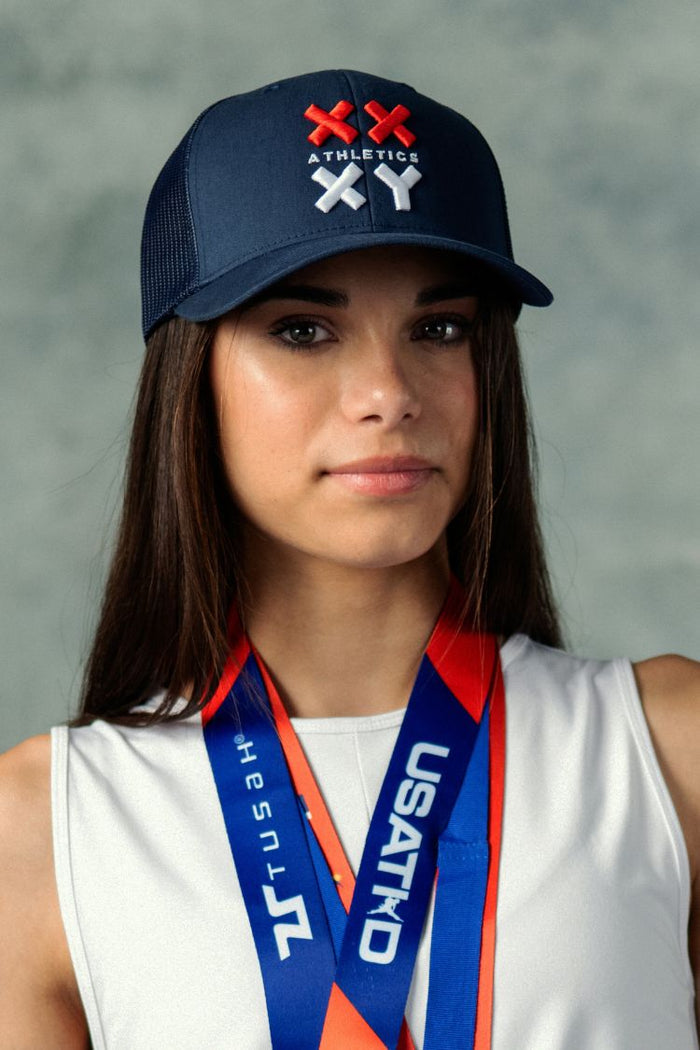 Woman wearing a navy cap with logos and a USA Today sports medal around her neck against a gray background || navy with red & white logo