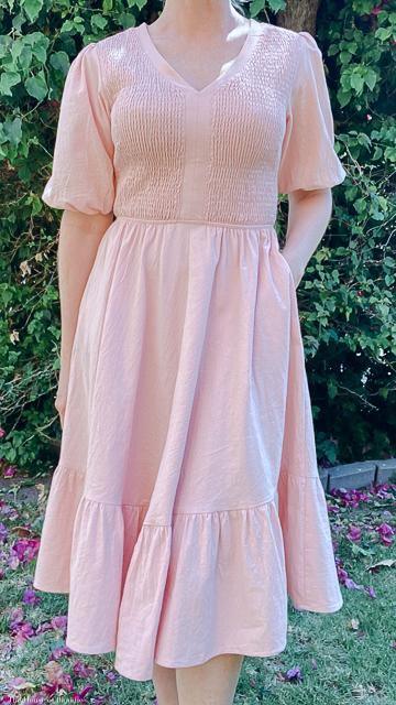 Woman wearing a pink smocked knee-length dress with puff sleeves, standing outdoors by greenery