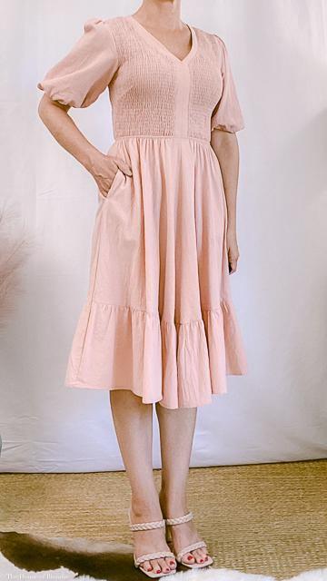 Woman wearing a pastel pink smocked knee-length dress with puff sleeves and ruffled hem in a minimalist setting