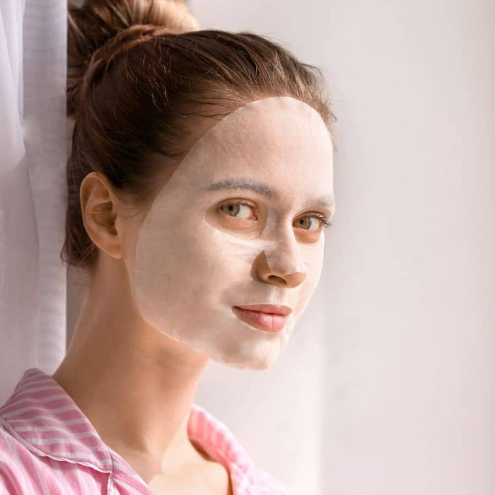 Young woman with a hydrating sheet mask applied, wearing a pink striped shirt, showcasing self-care and skincare wellness with Moodmask - The good fight | Patchology.