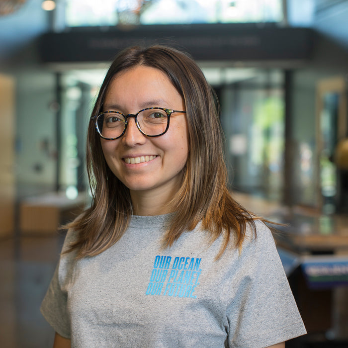 A smiling woman with glasses and shoulder-length brown hair wearing a heather gray t-shirt with a light blue graphic that reads 'OUR OCEAN OUR FUTURE' on the chest, standing in a brightly lit indoor environment.