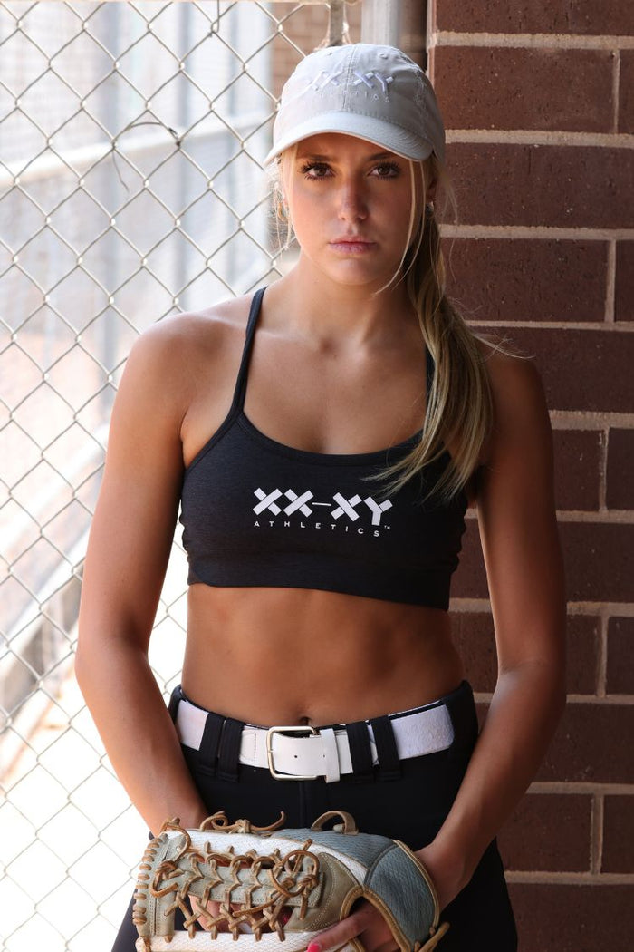 Woman wearing a black sports bra with 'XXXY Athletics' branding, holding a baseball glove against a brick wall.