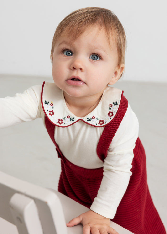 Baby wearing red knitted suspenders and white Christmas blouse with collar and floral embroidery