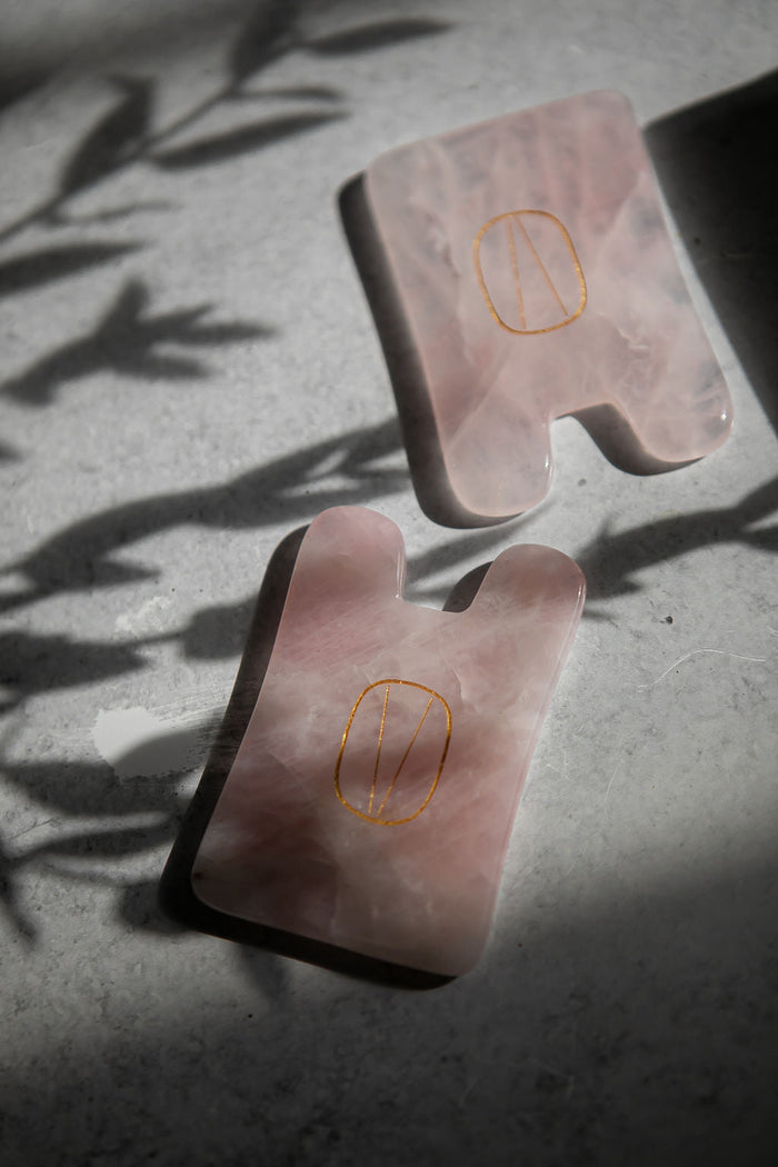 Two polished rose quartz facial massage tools engraved with a gold symbol, resting on a textured surface with soft natural shadows, Rose Quartz Gua Sha | Vie Healing.